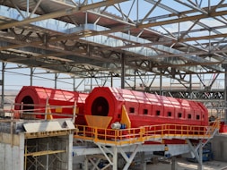 A large industrial facility featuring two prominent red cylindrical machines set within a metal framework. The machines are elevated and supported by a concrete structure, with yellow railings and metal scaffolding visible. The roof is made of metal beams in a grid pattern, partially open to the sky.