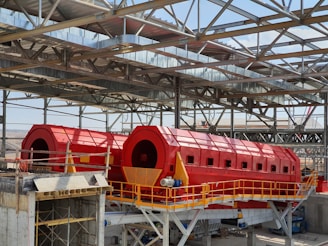 A large industrial facility featuring two prominent red cylindrical machines set within a metal framework. The machines are elevated and supported by a concrete structure, with yellow railings and metal scaffolding visible. The roof is made of metal beams in a grid pattern, partially open to the sky.