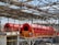 A large industrial facility featuring two prominent red cylindrical machines set within a metal framework. The machines are elevated and supported by a concrete structure, with yellow railings and metal scaffolding visible. The roof is made of metal beams in a grid pattern, partially open to the sky.
