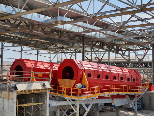 A large industrial facility featuring two prominent red cylindrical machines set within a metal framework. The machines are elevated and supported by a concrete structure, with yellow railings and metal scaffolding visible. The roof is made of metal beams in a grid pattern, partially open to the sky.