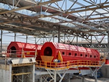 A large industrial facility featuring two prominent red cylindrical machines set within a metal framework. The machines are elevated and supported by a concrete structure, with yellow railings and metal scaffolding visible. The roof is made of metal beams in a grid pattern, partially open to the sky.