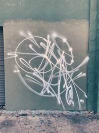 White abstract graffiti with looping lines and dots is painted on a weathered green wall. The rough texture of the wall complements the dynamic and flowing style of the graffiti. Below, the pavement is speckled with paint splatters, adding to the urban feel.