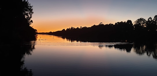 A tranquil lake at sunset, with calm water reflecting the silhouette of surrounding trees. The sky transitions from deep blue to vibrant orange near the horizon, creating a peaceful, serene atmosphere.