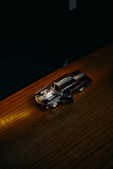 A small model car with an open door and a detailed engine under a spotlight on a dark wooden surface. The room around is dimly lit, creating a dramatic focus on the car.