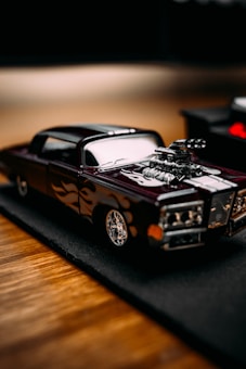 A detailed miniature model car with a sleek design and custom flame decals on the sides, featuring a prominent engine that sticks out of the hood. The car sits on a black mat, which contrasts with the wooden surface beneath.