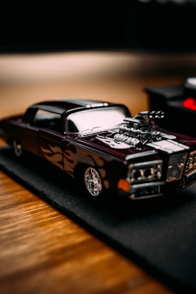 A detailed miniature model car with a sleek design and custom flame decals on the sides, featuring a prominent engine that sticks out of the hood. The car sits on a black mat, which contrasts with the wooden surface beneath.