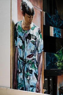 A mural painted on the side of a building depicting a person with short hair wearing a camouflage-patterned outfit, looking downward with a contemplative expression. The artwork stretches from the top to the bottom of the building wall.