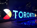 Large, illuminated letters spell out 'TORONTO' against a dark cityscape backdrop. The design includes a circular color wheel with sections in red, white, black, and yellow. Reflections appear on the wet ground, and a maple leaf at the end of the letters ties into a Canadian theme.