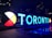 Large, illuminated letters spell out 'TORONTO' against a dark cityscape backdrop. The design includes a circular color wheel with sections in red, white, black, and yellow. Reflections appear on the wet ground, and a maple leaf at the end of the letters ties into a Canadian theme.
