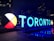 Large, illuminated letters spell out 'TORONTO' against a dark cityscape backdrop. The design includes a circular color wheel with sections in red, white, black, and yellow. Reflections appear on the wet ground, and a maple leaf at the end of the letters ties into a Canadian theme.