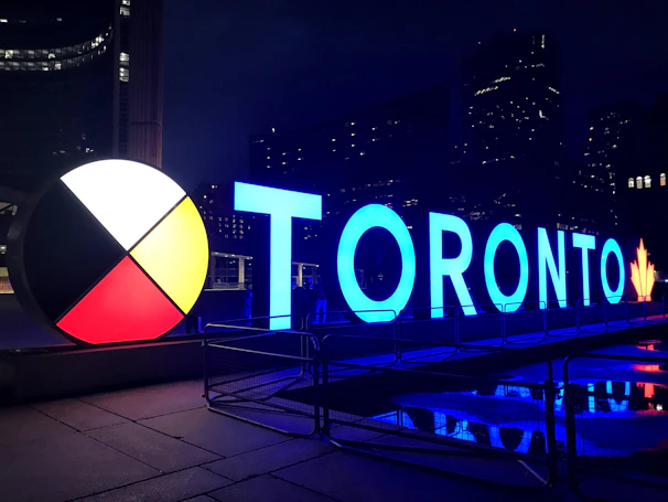 Large, illuminated letters spell out 'TORONTO' against a dark cityscape backdrop. The design includes a circular color wheel with sections in red, white, black, and yellow. Reflections appear on the wet ground, and a maple leaf at the end of the letters ties into a Canadian theme.