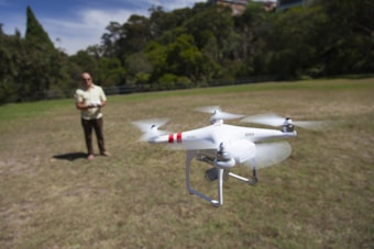 A person is operating a white quadcopter drone in a grassy area, surrounded by trees in the background. The drone is in flight, showing its propellers in motion, while the person stands in the distance holding a remote control.