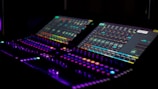A dimly lit control panel featuring two electronic screens with various colorful graphs and icons, illuminated buttons, and dials. The atmosphere is high-tech and futuristic, suggesting an audio or visual mixing setup.
