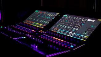 A dimly lit control panel featuring two electronic screens with various colorful graphs and icons, illuminated buttons, and dials. The atmosphere is high-tech and futuristic, suggesting an audio or visual mixing setup.