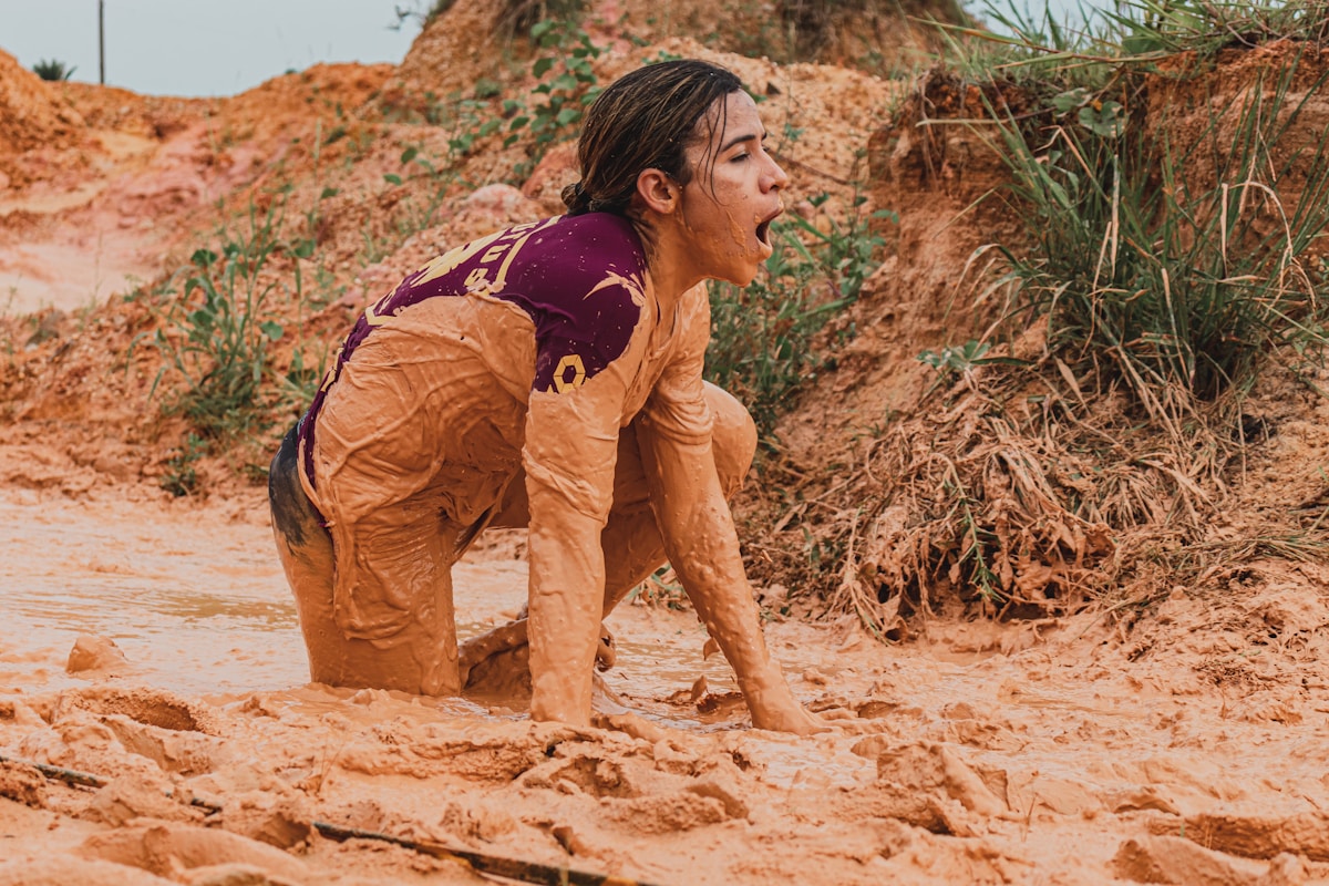 A dark haired woman crawling out of the mud.  She is wearing a wine colored shirt and her body is covered in mud.  Behind her is an embankment with more mud and sparse foliage.
