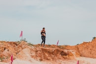 A person in athletic gear is walking on a dirt path surrounded by rugged terrain and marked with pink flags. The sky is overcast, and the ground is a mix of sand and clay.
