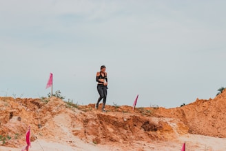 A person in athletic gear is walking on a dirt path surrounded by rugged terrain and marked with pink flags. The sky is overcast, and the ground is a mix of sand and clay.