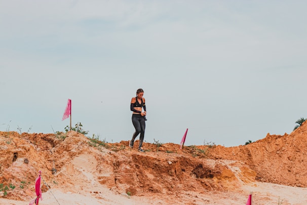 A person in athletic gear is walking on a dirt path surrounded by rugged terrain and marked with pink flags. The sky is overcast, and the ground is a mix of sand and clay.