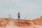 A person in athletic gear is walking on a dirt path surrounded by rugged terrain and marked with pink flags. The sky is overcast, and the ground is a mix of sand and clay.