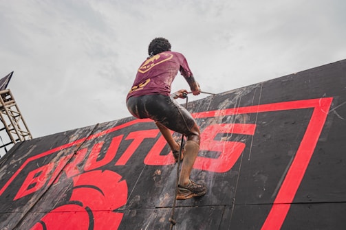 A person is climbing a slippery, inclined wall using a rope. The wall features the bold text 'BRUTUS' in red, and the climber is wearing a maroon shirt and black shorts, both covered in mud.