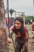 A person is participating in an outdoor obstacle course, grabbing onto a rope with determination. Their clothing is covered in mud, indicating a challenging and intense competition environment. In the background, there are palm trees and obstacle structures, contributing to the rugged and adventurous setting.