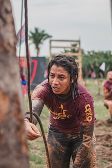 A person is participating in an outdoor obstacle course, grabbing onto a rope with determination. Their clothing is covered in mud, indicating a challenging and intense competition environment. In the background, there are palm trees and obstacle structures, contributing to the rugged and adventurous setting.