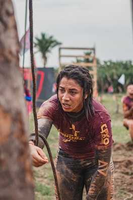 A person is participating in an outdoor obstacle course, grabbing onto a rope with determination. Their clothing is covered in mud, indicating a challenging and intense competition environment. In the background, there are palm trees and obstacle structures, contributing to the rugged and adventurous setting.