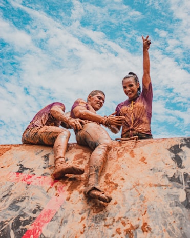 Runners climbing a muddy obstacle during the Heracles Race under a bright sky.
