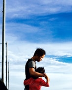 A man embraces a child, providing a sense of comfort and protection against the backdrop of a blue sky with scattered clouds. The child is wearing a red and black striped shirt, and the man is dressed in casual attire. A metal pole is visible on the left side of the image.
