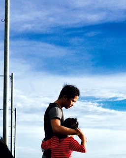 A man embraces a child, providing a sense of comfort and protection against the backdrop of a blue sky with scattered clouds. The child is wearing a red and black striped shirt, and the man is dressed in casual attire. A metal pole is visible on the left side of the image.