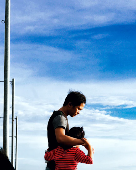 A man embraces a child, providing a sense of comfort and protection against the backdrop of a blue sky with scattered clouds. The child is wearing a red and black striped shirt, and the man is dressed in casual attire. A metal pole is visible on the left side of the image.