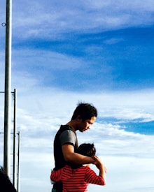 A man embraces a child, providing a sense of comfort and protection against the backdrop of a blue sky with scattered clouds. The child is wearing a red and black striped shirt, and the man is dressed in casual attire. A metal pole is visible on the left side of the image.