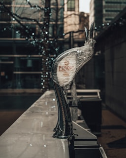 A beer tap wrapped in protective plastic is positioned on an outdoor bar counter. The background features a blurry urban setting with trees adorned by small, sparkling lights. The setting appears to be in a metropolitan area with tall buildings visible.