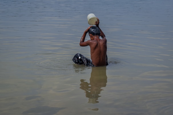 A person is partially submerged in water, pouring water over their head with a plastic container. The calm water reflects their image, and the environment appears serene.