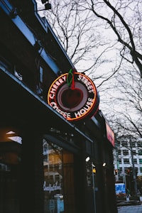 A neon sign in the shape of a cherry hangs outside a building, displaying the name 'Cherry Street Coffee House'. The sign is illuminated against a backdrop of bare tree branches and a cloudy sky. Part of the brick building's exterior is visible, along with a window displaying the words 'Breakfast Lunch'. Urban street elements like a road and distant buildings are partially visible.