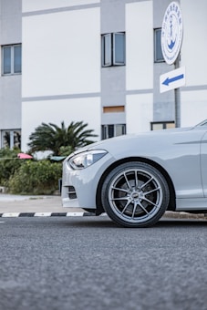 A silver car is parked on a road next to a building with a sign featuring an anchor logo and Chinese text. The car's front wheel and headlight are prominently visible, with a background of greenery and a modern structure.