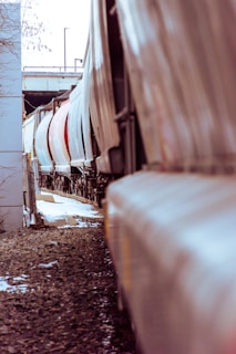 A row of freight train cars aligned on a railway track, viewed from a low angle. The cars are predominantly white with some red ones, stretching into the distance with a slight curve. The ground is covered in gravel, and there are patches of snow visible. Metal fencing and concrete structures can be seen on the left side, with an overpass visible in the background.