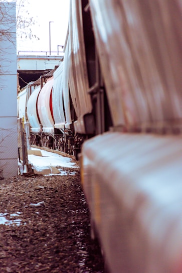 A row of freight train cars aligned on a railway track, viewed from a low angle. The cars are predominantly white with some red ones, stretching into the distance with a slight curve. The ground is covered in gravel, and there are patches of snow visible. Metal fencing and concrete structures can be seen on the left side, with an overpass visible in the background.