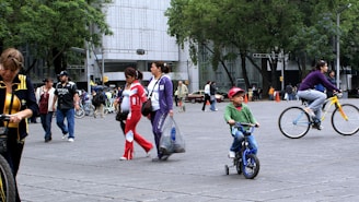 A busy urban street scene with people walking and cycling. In the foreground, a young child riding a small bicycle is wearing a red cap, while a woman nearby is riding a bicycle with yellow rims. Several pedestrians are dressed in varied colorful outfits, carrying bags and walking in different directions. The background features tall trees and a large modern building, indicating a city environment.