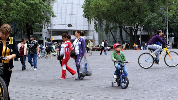 A busy urban street scene with people walking and cycling. In the foreground, a young child riding a small bicycle is wearing a red cap, while a woman nearby is riding a bicycle with yellow rims. Several pedestrians are dressed in varied colorful outfits, carrying bags and walking in different directions. The background features tall trees and a large modern building, indicating a city environment.