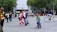 A busy urban street scene with people walking and cycling. In the foreground, a young child riding a small bicycle is wearing a red cap, while a woman nearby is riding a bicycle with yellow rims. Several pedestrians are dressed in varied colorful outfits, carrying bags and walking in different directions. The background features tall trees and a large modern building, indicating a city environment.