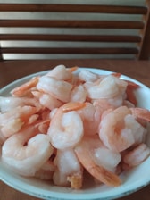 A bowl filled with peeled and cooked shrimp is placed on a wooden table with a chair visible in the background. The shrimp appear to have a light pink hue and are slightly glistening, suggesting freshness.