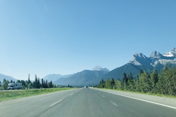 A clear roadway stretching into the distance with scenic mountains in the background. On the right side, lush green trees line the highway, while a recreational vehicle travels on the left, adding to the sense of freedom and exploration.