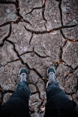 A pair of rugged hiking boots stand on dry, cracked ground, suggesting a harsh and arid environment. The texture of the earth shows deep fissures, indicating drought conditions.