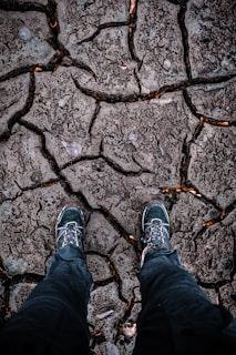 A pair of rugged hiking boots stand on dry, cracked ground, suggesting a harsh and arid environment. The texture of the earth shows deep fissures, indicating drought conditions.