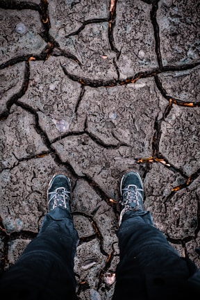 A pair of rugged hiking boots stand on dry, cracked ground, suggesting a harsh and arid environment. The texture of the earth shows deep fissures, indicating drought conditions.