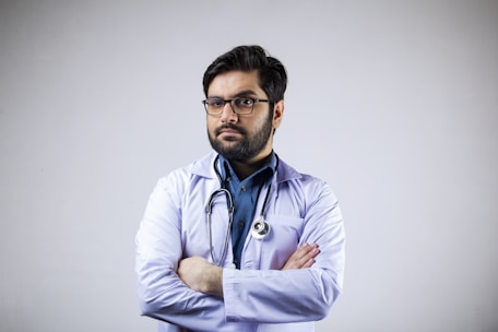 A man with a beard and glasses is wearing a white coat and a stethoscope around his neck. His arms are crossed, and he appears confident and professional. The background is plain and light-colored.