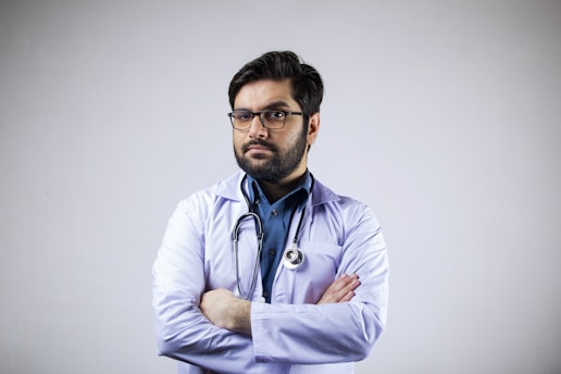 A man with a beard and glasses is wearing a white coat and a stethoscope around his neck. His arms are crossed, and he appears confident and professional. The background is plain and light-colored.