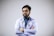 A man with a beard and glasses is wearing a white coat and a stethoscope around his neck. His arms are crossed, and he appears confident and professional. The background is plain and light-colored.