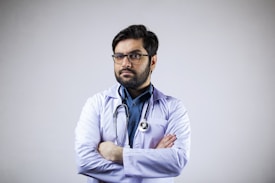 A man with a beard and glasses is wearing a white coat and a stethoscope around his neck. His arms are crossed, and he appears confident and professional. The background is plain and light-colored.
